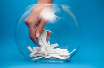 Una mano introduciéndose en un recipiente de cristal transparente lleno de papeletas blancas para realizar un sorteo aleatorio, representando el marco de las propiedades de la probabilidad